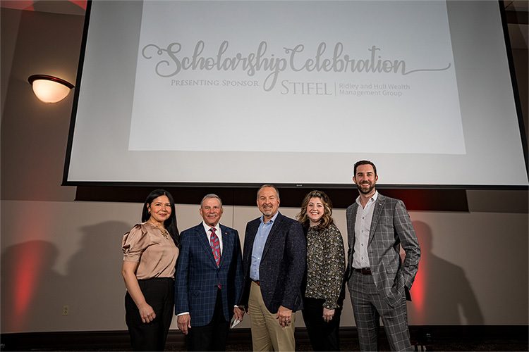 From left to right: Maria O'Neill, John W. Ridley, Derek Hull, Linsay Tambling, Drew Martin standing in front of project sign that reads; Scholarship Celebration Presenting Sponsor Stifel | Ridley and Hull Wealth Management Group
